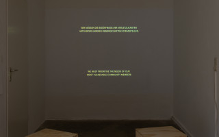 In der dunklen Raumnische wird Romily Alice Waldens „Notes rom the Underlands“ (2019) an die Wand projiziert. Es sind je zwei Zeilen hellgelbe Schrift oben und unten im Bild vor einem dunklen Hintergrund zu sehen. Davor stehen weiter im Vordergrund zwei h